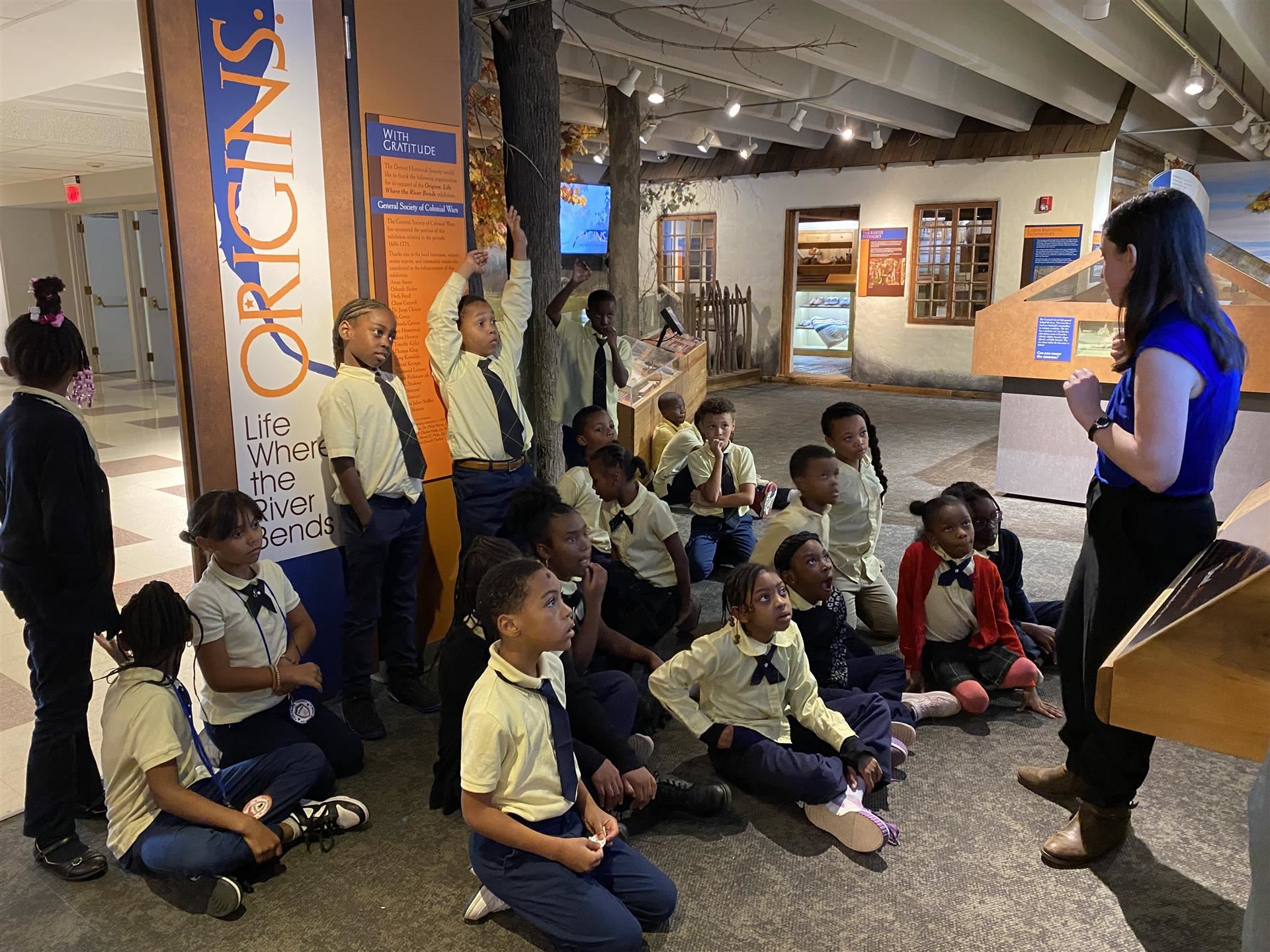 students sitting on the floor and raising their hands as they listen to a tour guide